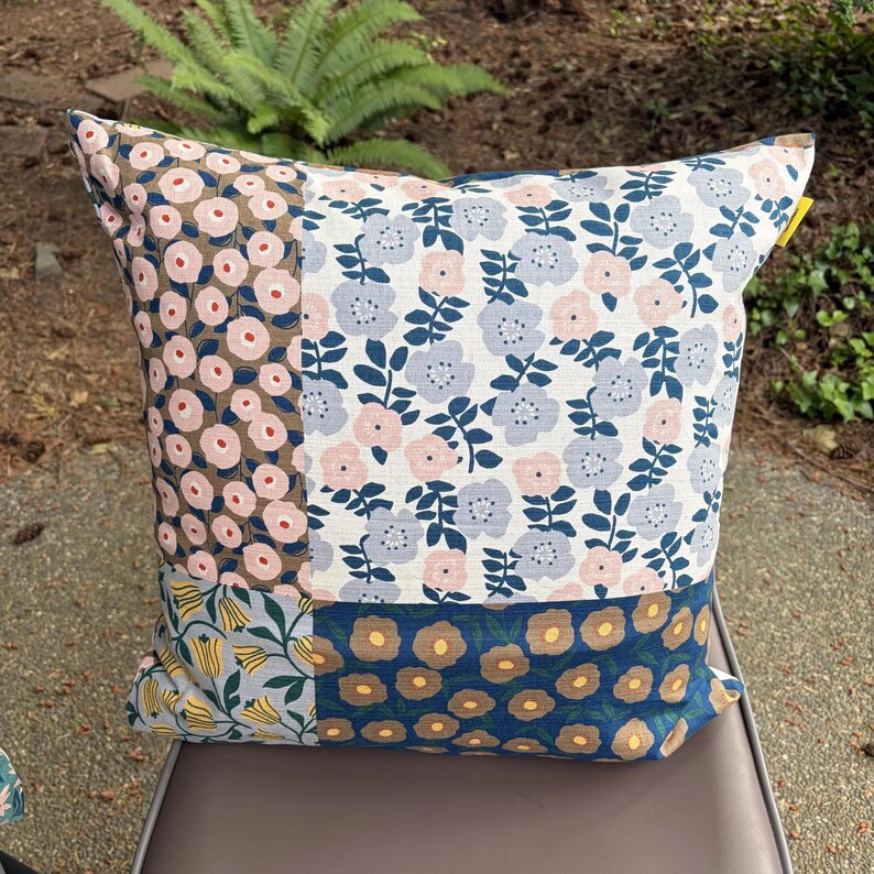 K&ouml;nnte beinhalten: Ein quadratisches Zierkissen mit einem Patchwork-Design aus vier verschiedenen Blumenmustern. Der Stoff zeigt eine Mischung aus hellrosa, staubblauen, gelben und dunkelbraunen Bl&uuml;tenmotiven auf wei&szlig;em, beigem und dunkelblauem Hintergrund mit gr&uuml;nen Stielen.