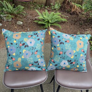 May include: Two square throw pillows featuring a teal background with a repeating floral pattern. The design includes stylized flowers in shades of tan, yellow, and blue, accented by dark brown stems and small orange circular motifs on each pillow.
