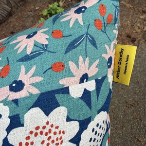 K&ouml;nnte beinhalten: Ein Kissenbezug aus blaugr&uuml;nem Stoff mit einem Blumenmuster aus hellrosa Bl&uuml;tenbl&auml;ttern, dunkelblauen Mitten und orangefarbenen Beeren. Ein gelbes Stoffetikett an der Seite zeigt den Text Atelier Dorothy Japanese Fabric in Schwarz.