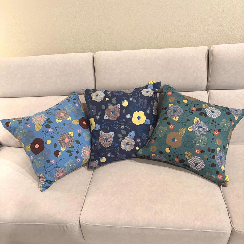 Puede incluir: Tres cojines cuadrados con un patr&oacute;n floral de flores estilizadas en tonos azul, amarillo, marr&oacute;n y blanco. Los cojines tienen fondos azul claro, azul marino oscuro y verde azulado, cada uno con una peque&ntilde;a etiqueta de tela amarilla.