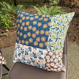 K&ouml;nnte beinhalten: Ein quadratisches Zierkissen mit einem Patchwork-Design aus vier verschiedenen floralen Stoffmustern. Die Abschnitte zeigen Blau mit beigen Blumen, Wei&szlig; mit gelben Tulpen, Wei&szlig; mit grauen und rosa Blumen sowie Beige mit rosa Blumen.