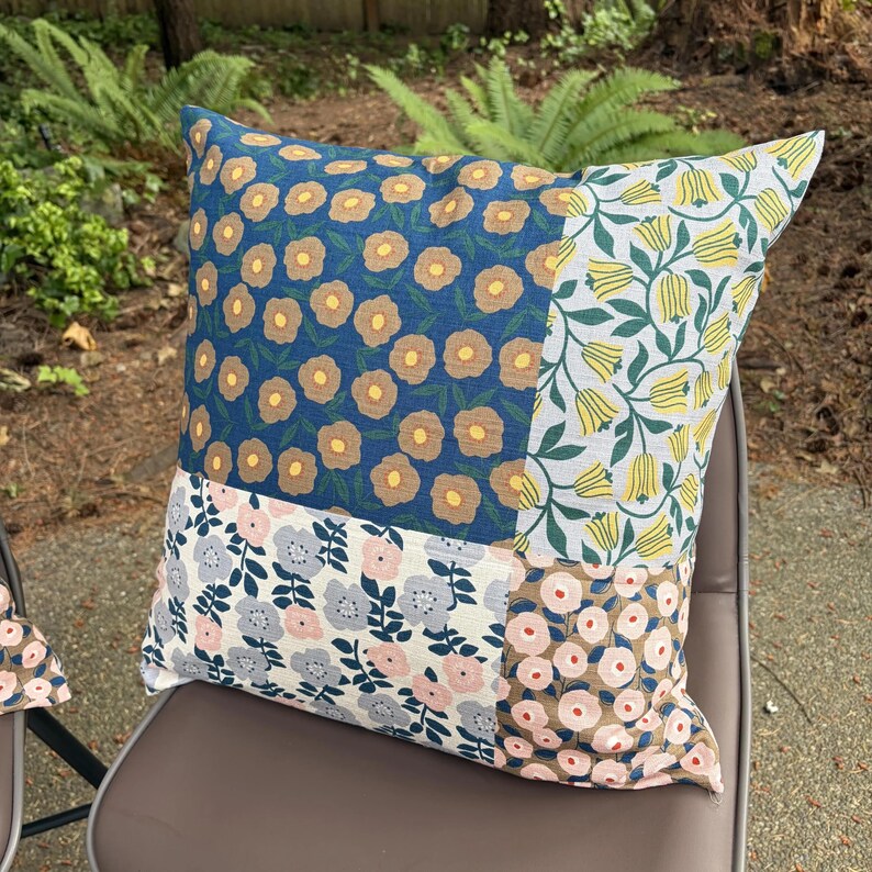 K&ouml;nnte beinhalten: Ein quadratisches Zierkissen mit einem Patchwork-Design aus vier verschiedenen floralen Stoffmustern. Die Abschnitte zeigen Blau mit beigen Blumen, Wei&szlig; mit gelben Tulpen, Wei&szlig; mit grauen und rosa Blumen sowie Beige mit rosa Blumen.