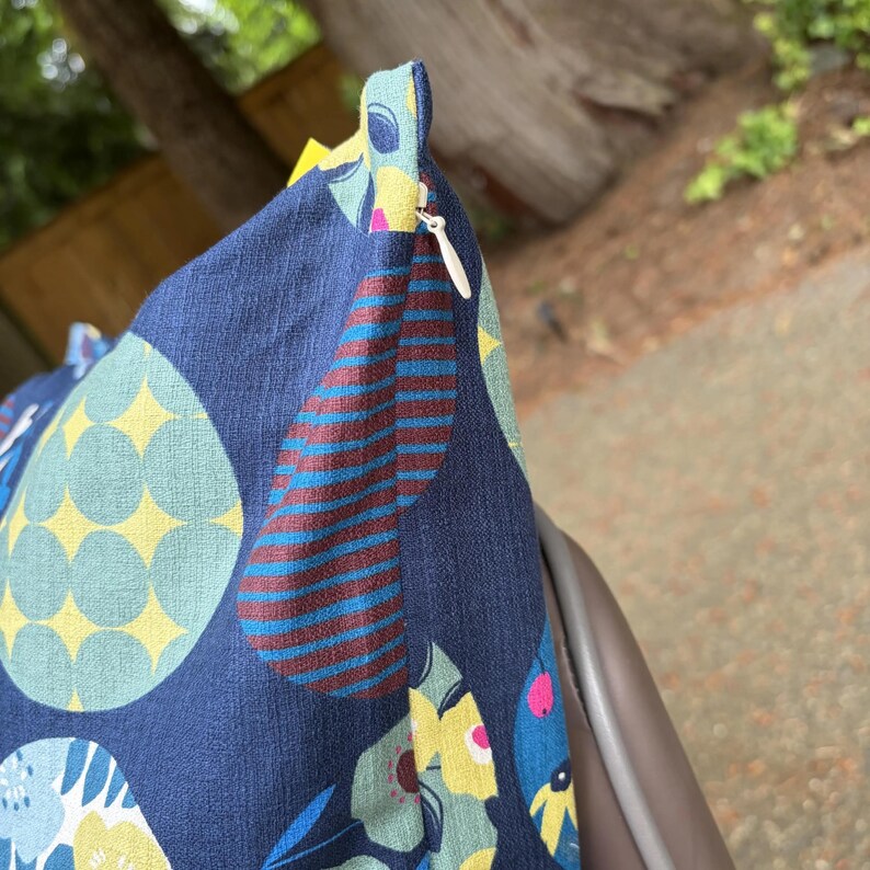 May include: A square throw pillow cover featuring a dark blue base with a multi-colored pattern of large circles, striped teardrop shapes, and floral motifs in yellow, light blue, and pink. A white zipper is visible along the side seam.