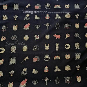Puede incluir: Una tela negra con un patrón repetitivo de pequeños iconos de inspiración japonesa, incluyendo máscaras, animales y objetos tradicionales. El texto "Cutting direction" con una flecha y "SozaiStudio" están impresos en la tela.
