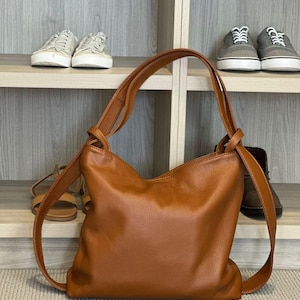 May include: A tan leather shoulder bag with a relaxed, unstructured shape. The bag features long straps and a simple design. The bag is placed on a beige carpet.