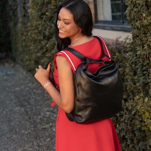 Könnte beinhalten: Ein schwarzer Lederrucksack mit runder Form wird über den Schultern getragen. Die Person trägt ein rotes Kleid mit weißen Streifen und Besatz. Der Hintergrund zeigt grünes Laub und ein Gebäude.