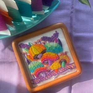 May include: A square, wooden-framed embroidery featuring colorful mushrooms. The mushrooms are embroidered with vibrant threads in shades of orange, yellow, green, and purple. A teal tray with spools of thread in various colors sits in the background.