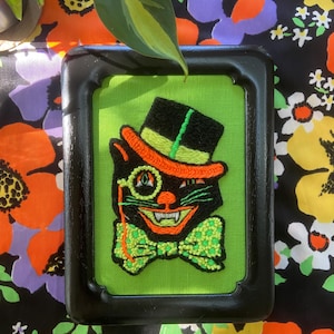 May include: A framed embroidered artwork featuring a black cat wearing a top hat, monocle, and bow tie. The cat has an orange hat and accents, a green bow tie, and a mischievous grin. The background is lime green, and the frame is black.
