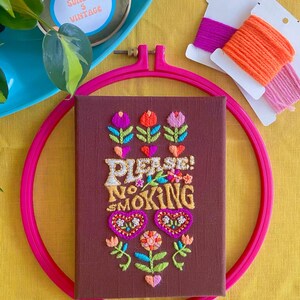 Peut inclure: Art de broderie sur un cercle avec un fond brun et le texte "PLEASE! NO SMOKING" en fil doré. Le texte est entouré de fleurs et de cœurs colorés.