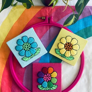 Op de afbeelding: Drie geborduurde bloemencanvas met verschillende gekleurde bloemen. De bloemen zijn blauw, geel en regenboogkleurig. De canvas worden getoond in een roze borduurring.