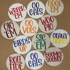 May include: A collection of twelve circular white fabric buttons featuring embroidered text in various colors. The text includes phrases such as WRECK EM, GO CATS, HOOK EM, GIG EM, GO VOLS, WOO PIG, BIRDS UP, SIC EM, GEAUX TIGERS, ROLL TIDE, and BOOMER SOONER.
