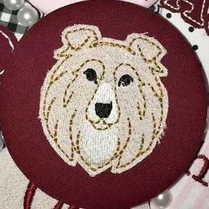May include: A circular burgundy fabric badge featuring an embroidered cream-colored dog face with gold-toned thread outlines. The dog has black embroidered eyes and a black nose, with a textured stitch pattern defining the fur and facial features.