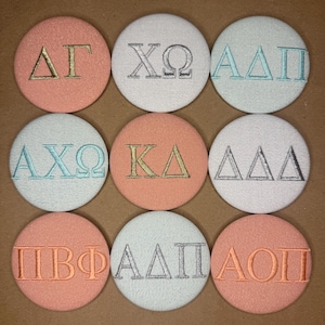 May include: Nine circular fabric buttons arranged in a grid, each featuring embroidered Greek letters. The buttons come in shades of peach, white, and light blue, with metallic gold, silver, and light blue thread used for the lettering.