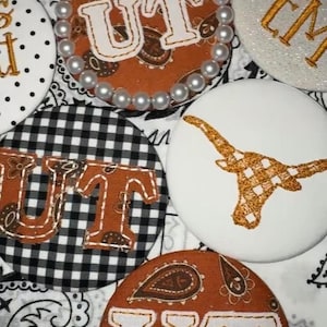 May include: A collection of circular fabric patches featuring various designs including the letters UT in white, a white longhorn steer silhouette, black and white gingham patterns, brown paisley prints, and white pearl bead embellishments.