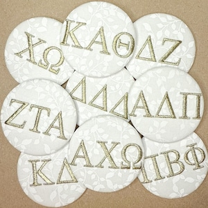 May include: A collection of round, white fabric coasters with gold embroidered Greek letters. The coasters feature a delicate leaf pattern and are arranged in a circular pattern. The Greek letters are in various combinations.