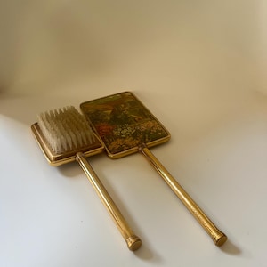 May include: A vintage vanity set comprising a brush and a hand mirror. Both have gold-toned metal frames and handles. The brush has a rectangular head with bristles, and the mirror has a rectangular face with a landscape scene.