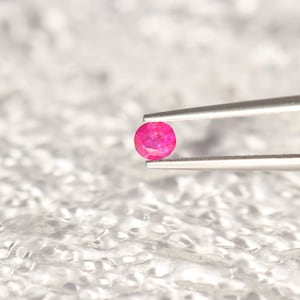 May include: A small, round, faceted gemstone with a vibrant pinkish-red hue held by a pair of silver-toned metal tweezers. The gemstone displays multiple cut surfaces that reflect light, showcasing its clarity and saturated color profile.