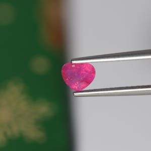 May include: A heart-shaped gemstone with a vibrant hot pink hue and a faceted surface texture. The stone is held securely between the two thin, metallic prongs of a pair of silver-toned tweezers, showcasing its distinct shape and bright colour.