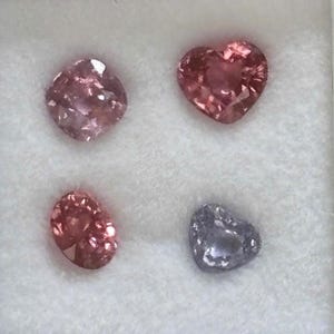 May include: Four faceted gemstones arranged in a square formation. The top left is a cushion-cut pink stone, the top right is a heart-cut pink stone, the bottom left is an oval-cut pink stone, and the bottom right is a heart-cut light purple stone.
