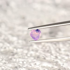 以下が含まれることがあります： 銀色の金属製ピンセットで保持された、小さなハート型の半透明の紫色の宝石。宝石は光を捉えるファセット加工された表面を持ち、全体にさまざまな紫と薄いラベンダーの色合いを見せています。