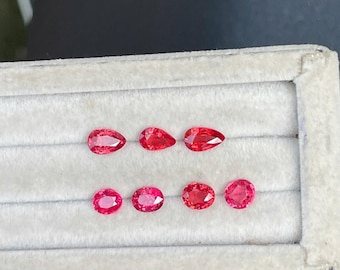 Puede incluir: Una colección de siete piedras preciosas facetadas sueltas en un tono rojo vibrante. La fila superior presenta tres piedras en forma de pera, mientras que la fila inferior contiene cuatro piedras ovaladas. Cada gema muestra un corte brillante.
