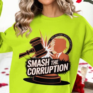 May include: A bright green sweatshirt featuring a graphic design of a gavel smashing a circle with the text "Smash the Corruption" and a silhouette of a person within the circle. The circle also includes the text "Asset Pelront Buet's Grottothee".