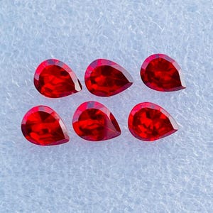 Pode incluir: Seis pedras preciosas facetadas em forma de pera de uma cor vermelha profunda e vibrante. Cada pedra apresenta uma extremidade pontiaguda e uma base arredondada com múltiplas facetas que refletem a luz, criando um aspeto brilhante e cintilante.
