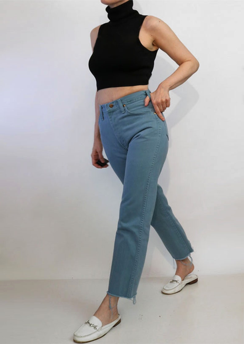 May include: A person wearing a black sleeveless turtleneck top and light blue jeans. The jeans have a raw hem and the person is wearing white loafers.