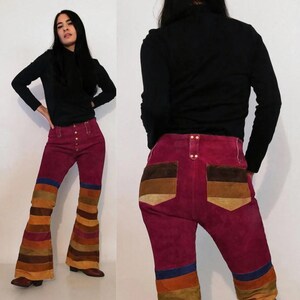 May include: High-waisted suede pants in a deep magenta hue featuring a flared leg design. The lower legs and back pockets are accented with horizontal stripes in shades of brown, tan, and blue. The pants include a front button-up closure.