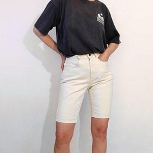 May include: A dark charcoal gray short-sleeve t-shirt with a small white graphic on the chest paired with off-white knee-length denim shorts featuring a button and zipper fly closure and a classic five-pocket design.