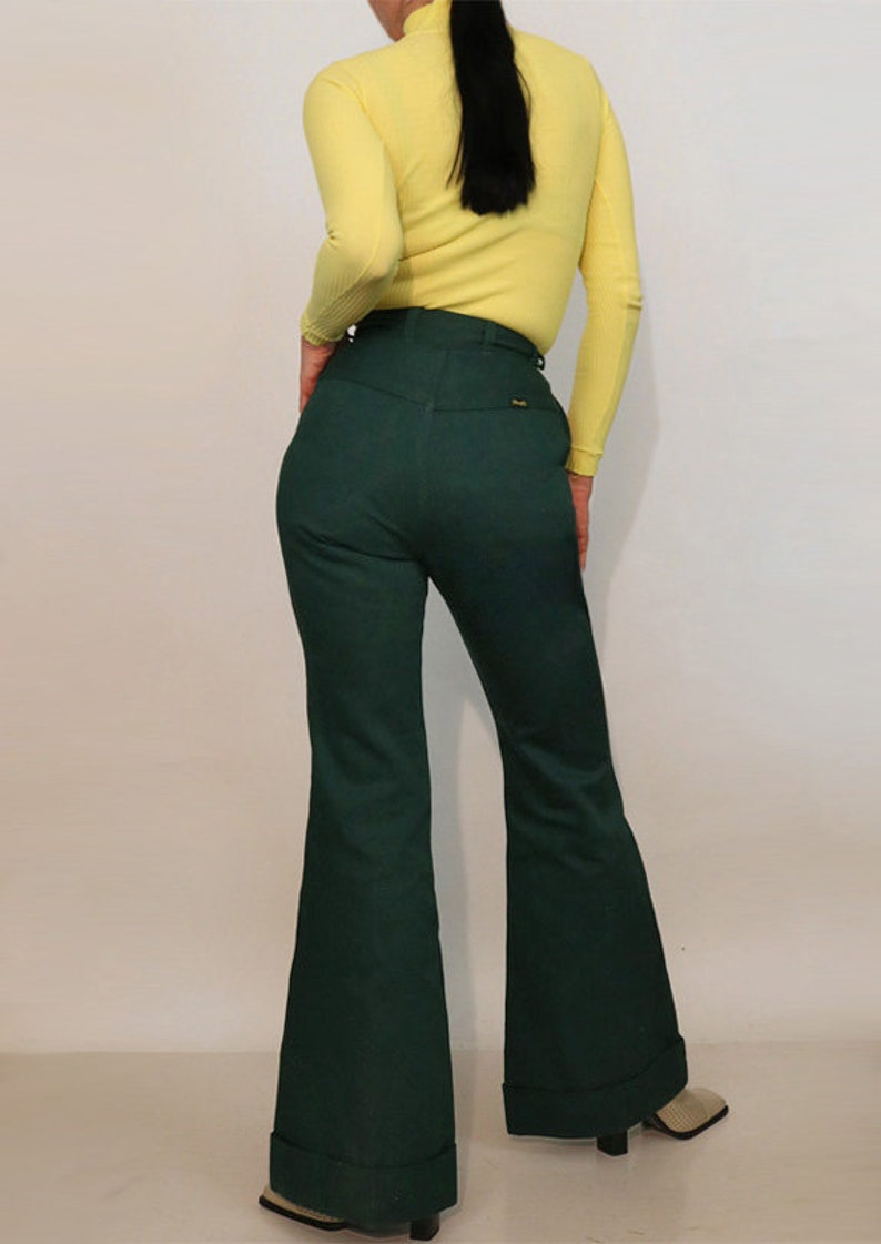 Puede incluir: Un par de pantalones de campana de pierna ancha, de cintura alta, de color verde oscuro con dobladillo vuelto. Los pantalones tienen un bolsillo trasero con una peque&ntilde;a etiqueta.