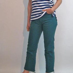 Puede incluir: Una camiseta a rayas azules y blancas y unos vaqueros verde azulado con dobladillo sin rematar. El conjunto se completa con sandalias negras. La ropa informal es adecuada para el uso diario.