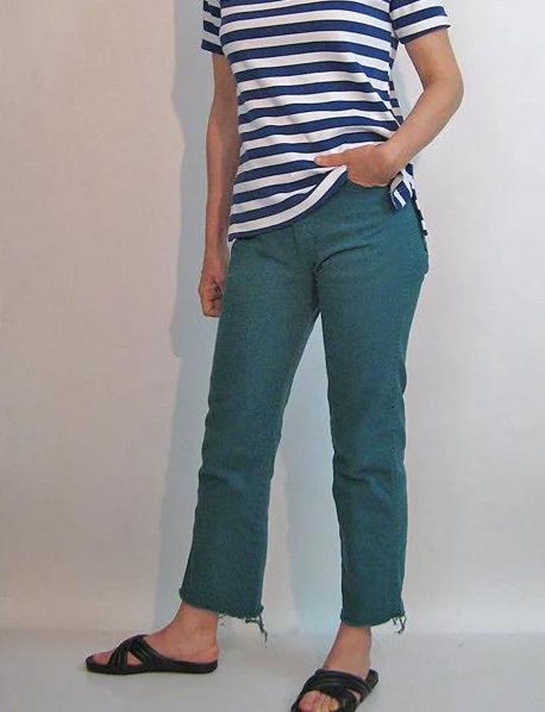 Puede incluir: Una camiseta a rayas azules y blancas y unos vaqueros verde azulado con dobladillo sin rematar. El conjunto se completa con sandalias negras. La ropa informal es adecuada para el uso diario.