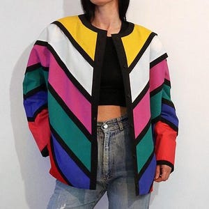 May include: A long-sleeved jacket featuring a bold, multi-colored chevron pattern in yellow, white, pink, teal, blue, and red. The garment has a round neckline, a black front button closure, and black trim separating each diagonal color stripe.