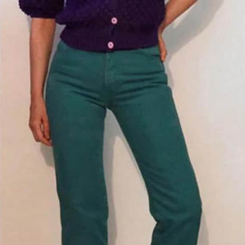 Puede incluir: Una persona con una rebeca morada con botones rosas y vaqueros verde azulado. La rebeca tiene mangas cortas. Los vaqueros son de color s&oacute;lido y tienen un corte recto.