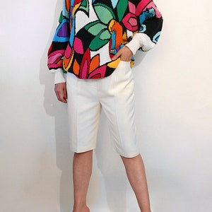 Puede incluir: Un su&eacute;ter blanco con un estampado floral abstracto de colores. El su&eacute;ter tiene mangas largas y un corte holgado. La modelo lleva pantalones cortos blancos y sandalias negras de tac&oacute;n.