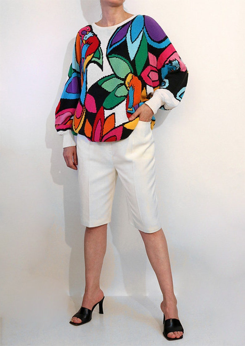 Puede incluir: Un su&eacute;ter blanco con un estampado floral abstracto de colores. El su&eacute;ter tiene mangas largas y un corte holgado. La modelo lleva pantalones cortos blancos y sandalias negras de tac&oacute;n.