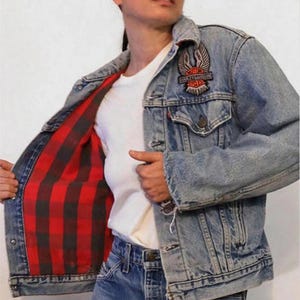 May include: A light blue denim jacket with a red and black checked lining. The jacket features a Harley Davidson logo patch on the left side of the chest.