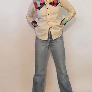 May include: A cream colored long-sleeved button-down shirt with a patchwork design on the yoke. The shirt has a pointed collar and a single button closure at the cuffs. The model is wearing light blue jeans and brown sandals.