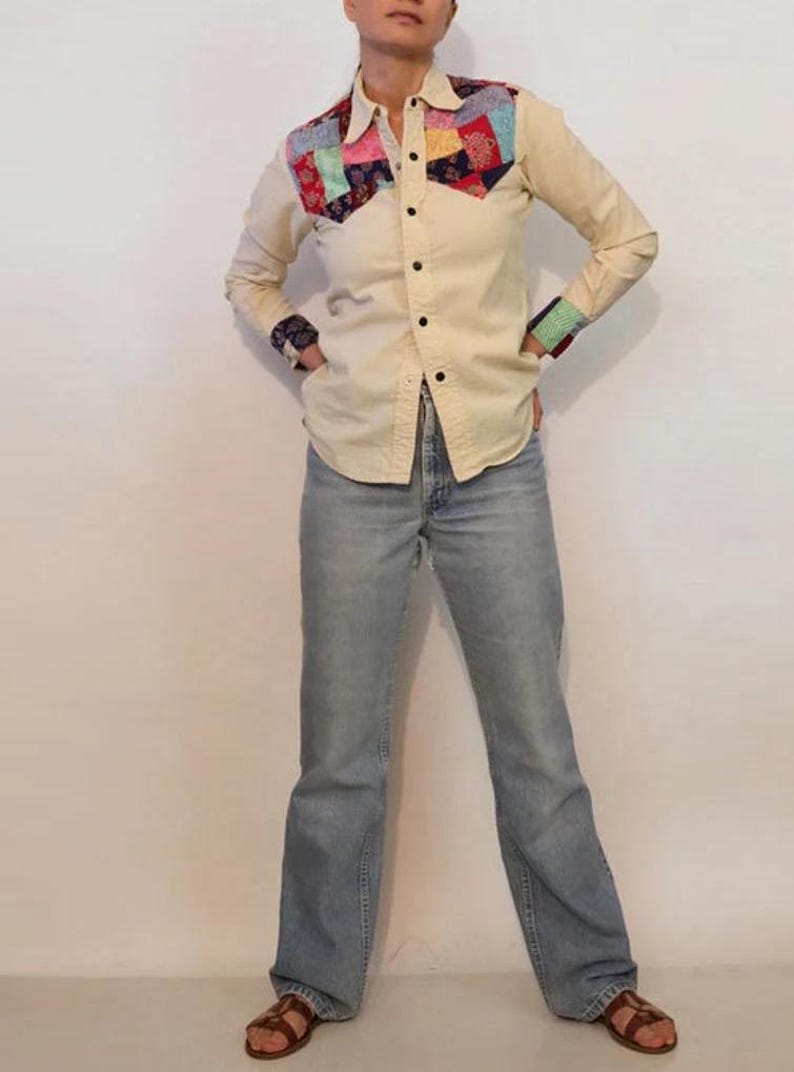 May include: A cream colored long-sleeved button-down shirt with a patchwork design on the yoke. The shirt has a pointed collar and a single button closure at the cuffs. The model is wearing light blue jeans and brown sandals.