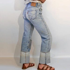May include: Light blue denim jeans with a distressed look, featuring a patched knee and large cuffs. The jeans have a faded appearance with visible wear and tear. Brown leather sandals complete the outfit.