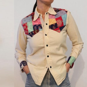 May include: A cream-coloured long-sleeved shirt with a patchwork design on the yoke. The patchwork is made of various fabrics in shades of red, blue, green, and pink.