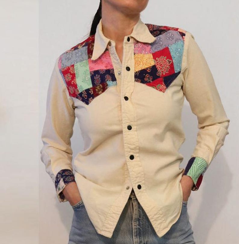 May include: A cream-colored long-sleeved shirt with a patchwork design on the yoke. The patchwork is made of various fabrics in shades of red, blue, green, and pink.