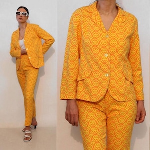 May include: A bright yellow and orange patterned two-piece suit consisting of a long-sleeve blazer with a notched lapel and three front buttons, paired with matching trousers. The fabric features a repeating geometric motif throughout the set.