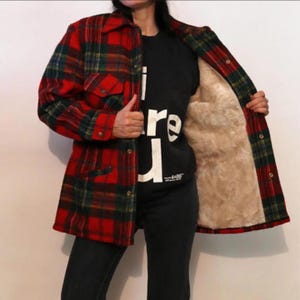 May include: A red, green and yellow plaid jacket with a tan faux fur lining. The jacket features a collared neckline, long sleeves and a button-front closure. It is worn over a black shirt with white text that reads "I are u".