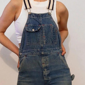 May include: Blue denim dungarees with white and black striped straps. The dungarees have a large front pocket and a button closure.