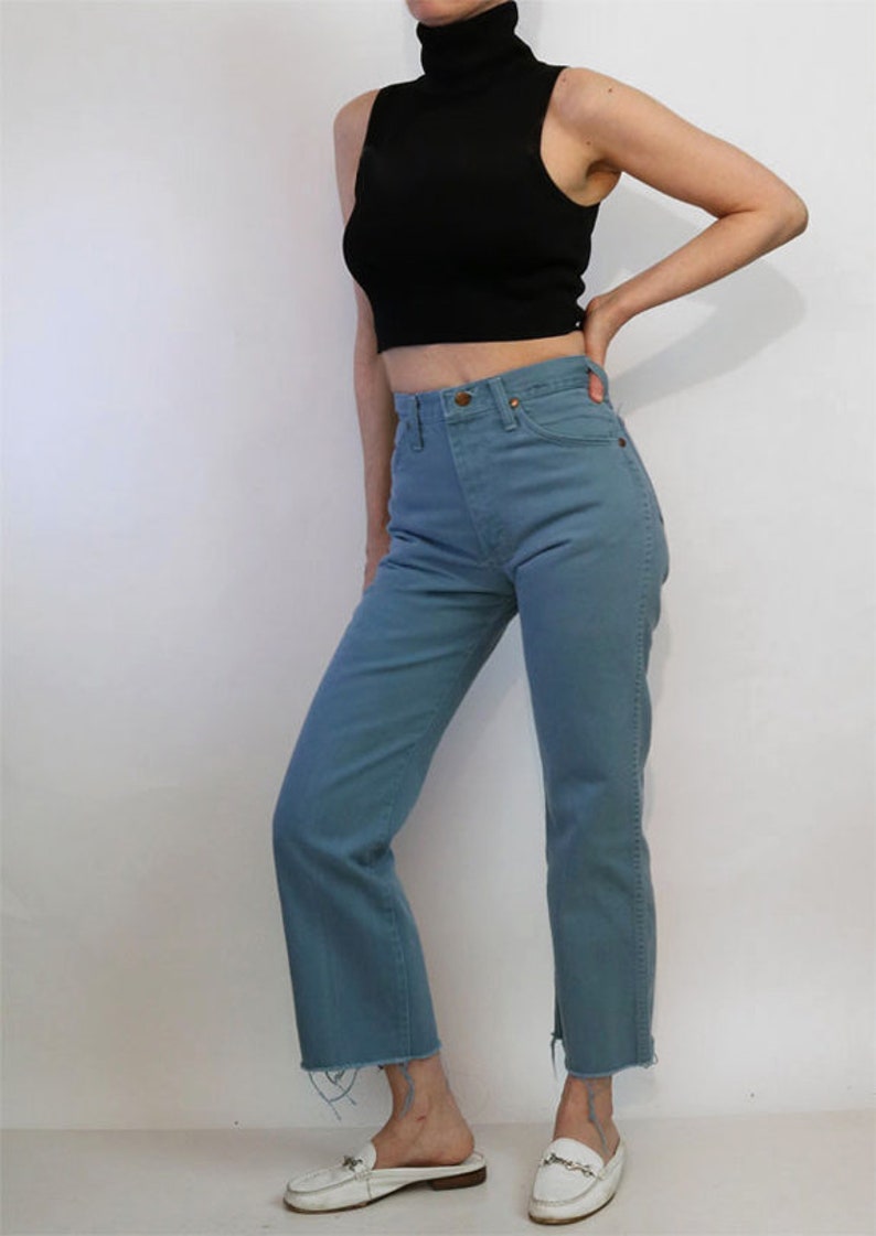 May include: A woman is wearing a black sleeveless turtleneck top and blue denim jeans with a raw hem. She is wearing white leather loafers.