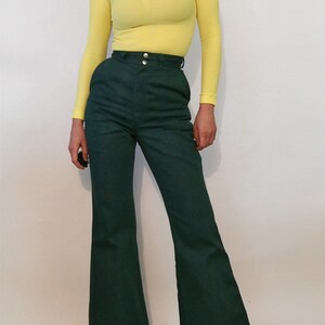 Puede incluir: Una mujer lleva un jersey de cuello alto amarillo brillante y pantalones de pierna ancha verde oscuro con cintura alta.