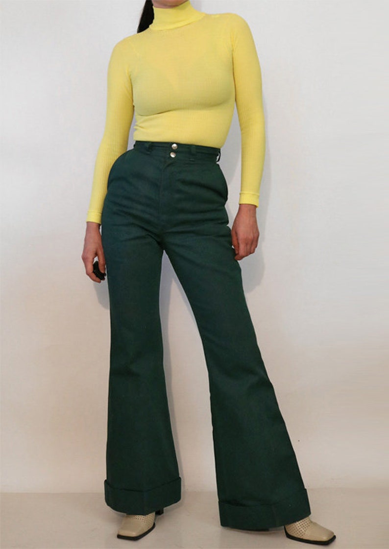 Puede incluir: Una mujer lleva un jersey de cuello alto amarillo brillante y pantalones de pierna ancha verde oscuro con cintura alta.