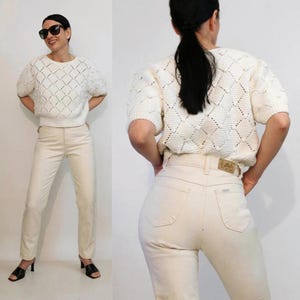 May include: A cream-coloured short-sleeve knit jumper featuring a diamond-patterned texture with small raised dots, paired with light beige denim jeans. The jeans have a tan leather patch on the back waistband with the brand name Lee printed on it.